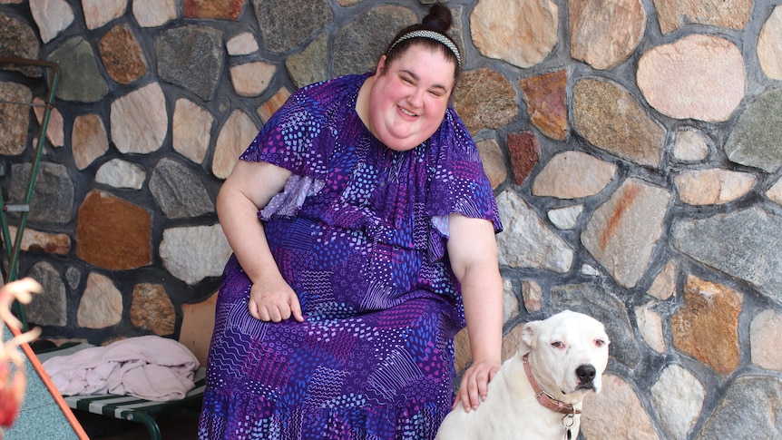 Woman in purple dress sitting on seat outside house with stone featuring patting cream coloured dog, doghouse in left corner.