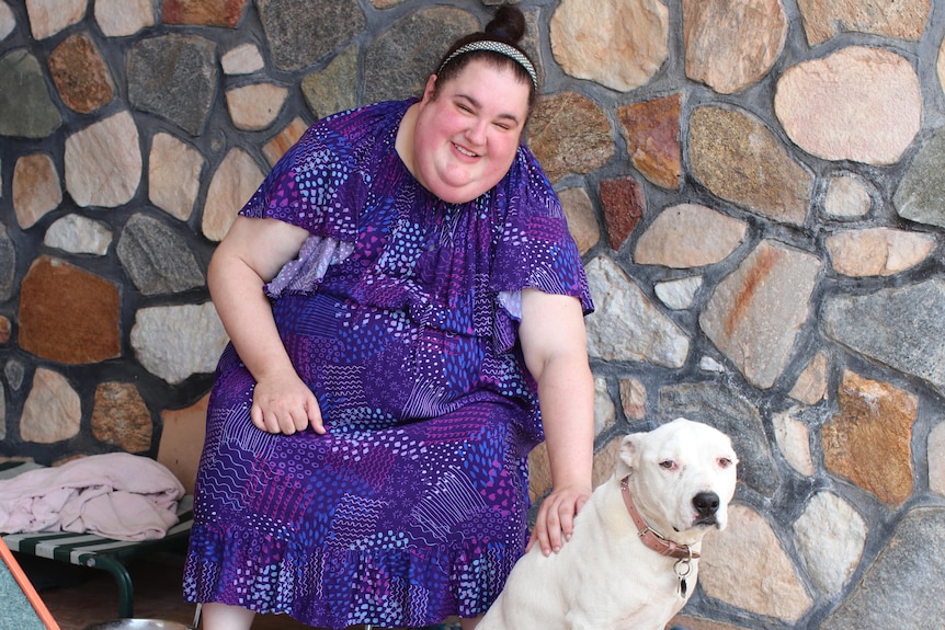 Woman in purple dress sitting on seat outside house with stone featuring patting cream coloured dog, doghouse in left corner.