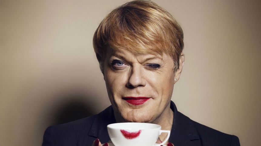 Eddie Izzard holds up a teacup with a lipstick mark on it and winks at the camera.