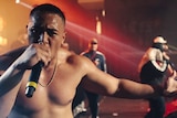 A shirtless man raps into a microphone onstage, with other rappers behind him. 