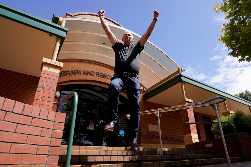 A man in jeans and a black t-shirt jumps with his arms raised