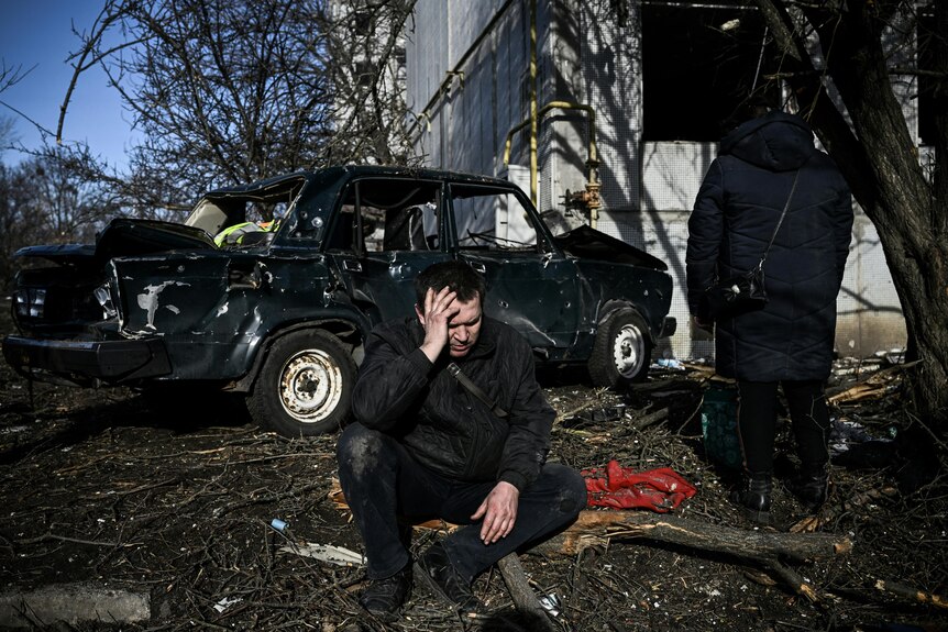 A man sits in front of a car covered in bullet holes
