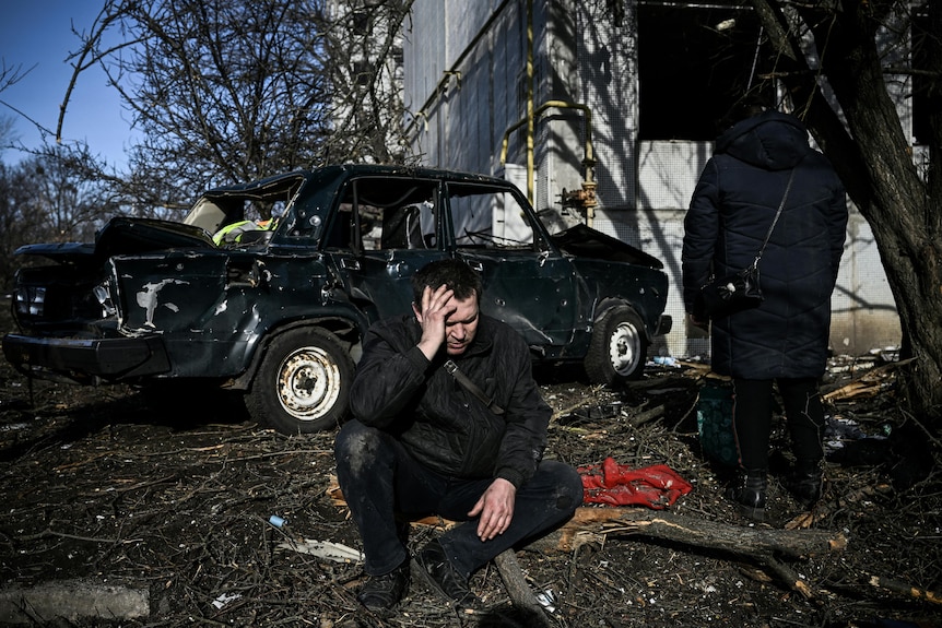 A man sits in front of a car covered in bullet holes
