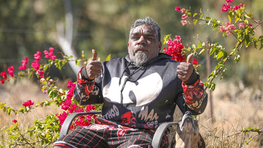 An Indigenous man poses with his thumbs up at the camera, sitting in front of a bougainvillea plant. A dog sits next to him.