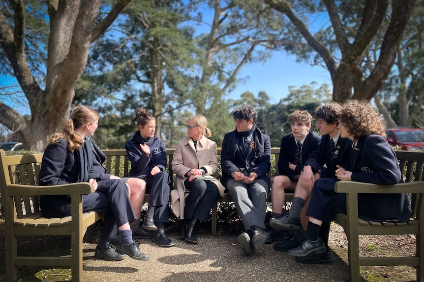 Emma Grant and seven year 11 students in formal school uniforms sit on a park bench outside and talk.
