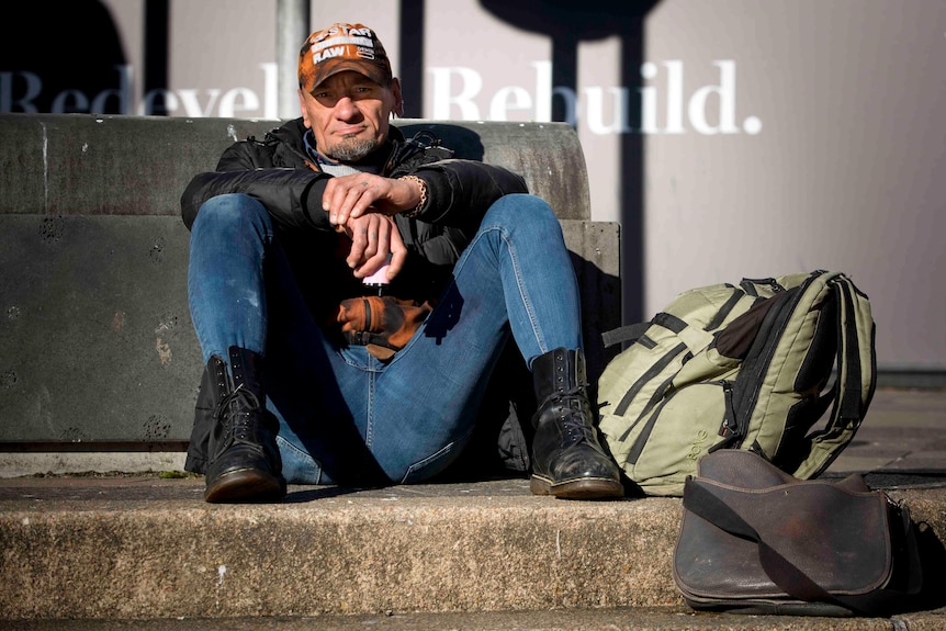 A man with blue jeans and a leather jacket sitting on concrete outside