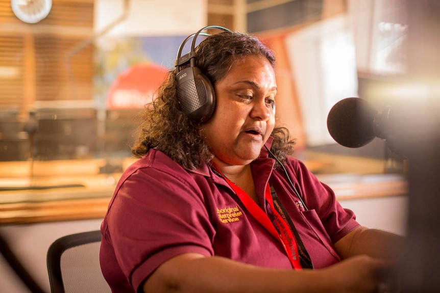 An Aboriginal woman reads into a microphone from a script in a radio broadcasting studio.