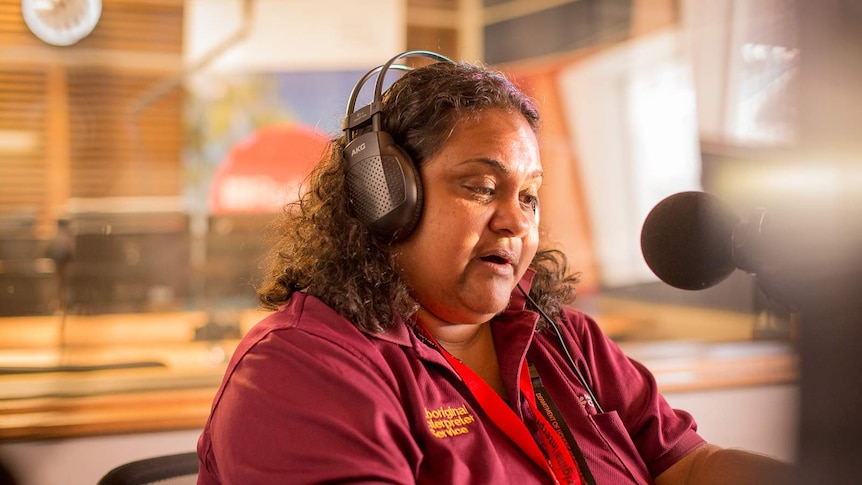 An Aboriginal woman reads into a microphone from a script in a radio broadcasting studio.