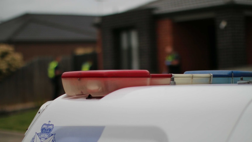 The back of a police van, with red and blue siren lights and the blue Victoria Police logo is shown in front of a house.