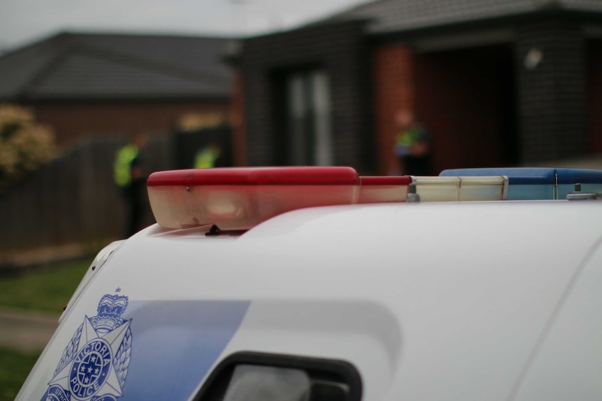 The back of a police van, with red and blue siren lights and the blue Victoria Police logo is shown in front of a house.