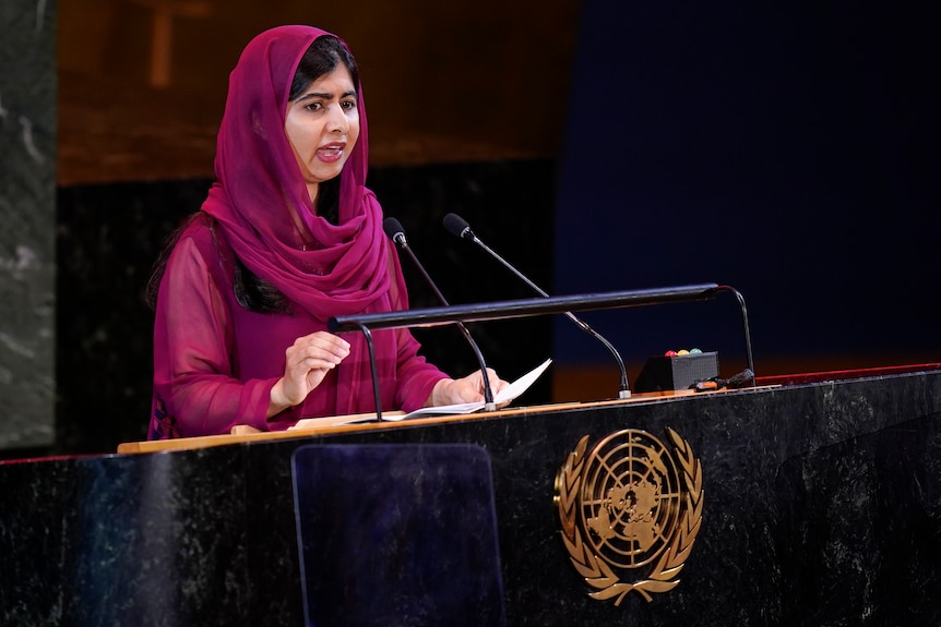 Malala Yousafzai, wearing pink, speaks at a lectern. 