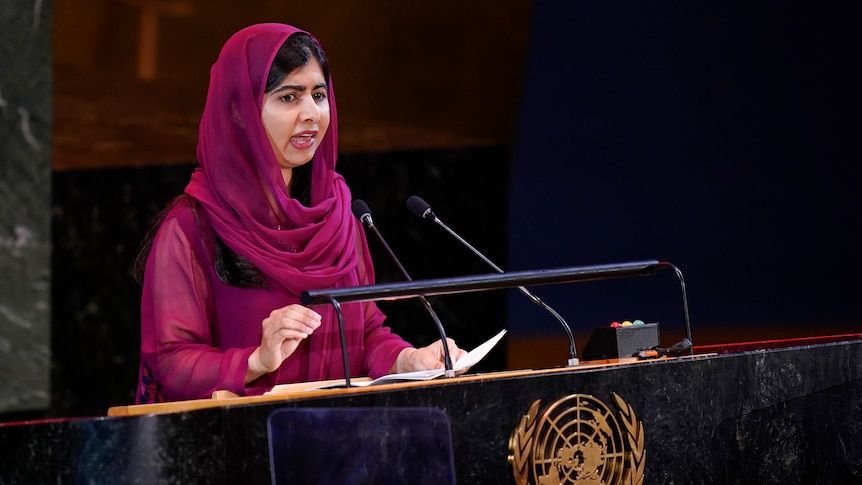 Malala Yousafzai, wearing pink, speaks at a lectern. 