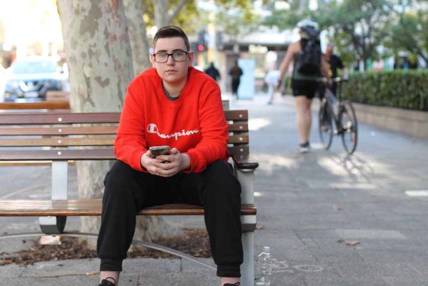 boy sitting on bench in city looking at camera with his phone in his hands