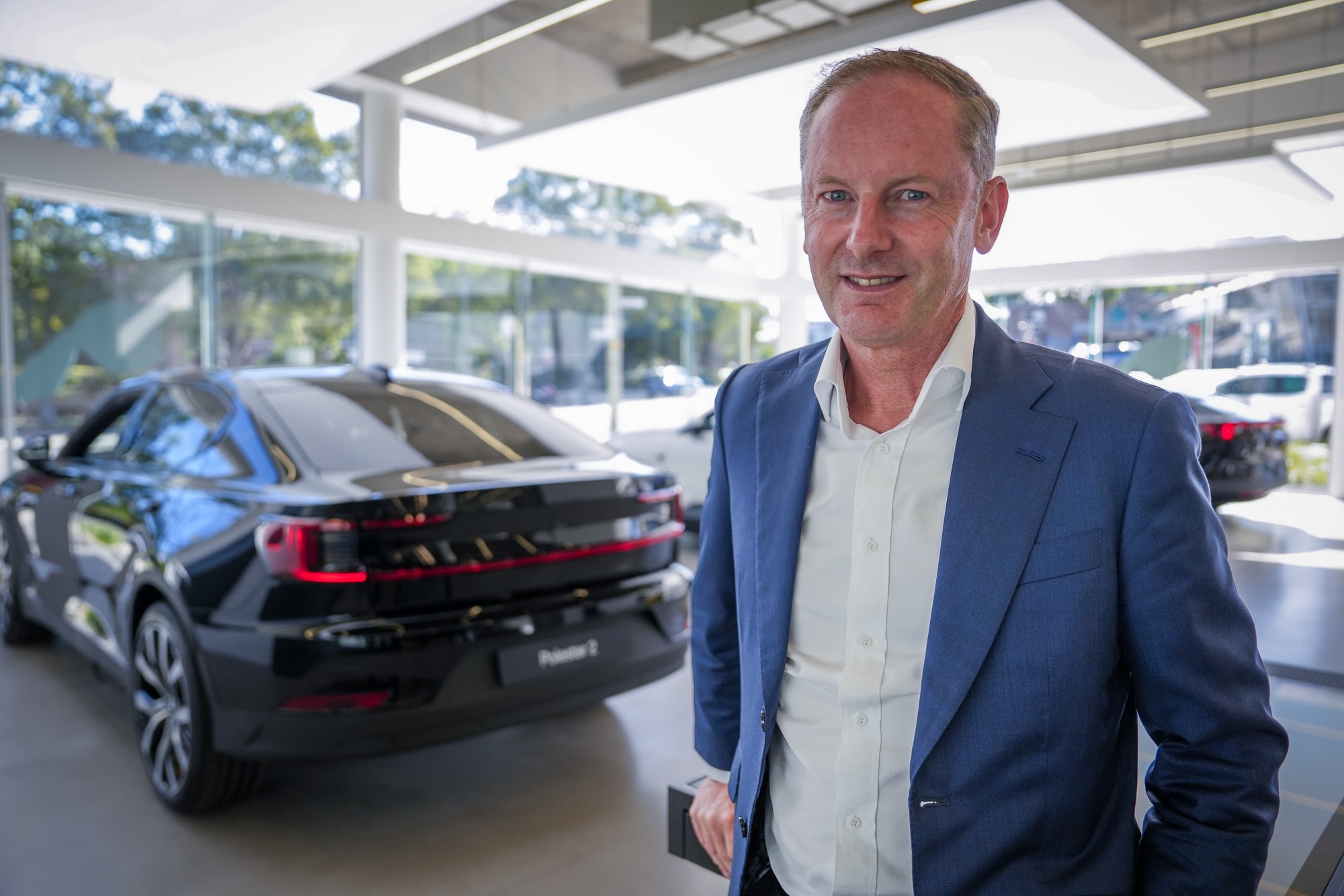 Scott Maynard stands in front of car, smiles, wears a blue suit, white shirt. Caucasian, receding hairline.