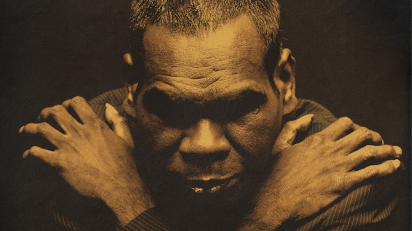 An shadowy portrait of Gamitj man and Yolgnu musician Gurrumul posing in a suit with his arms crossed his chest.