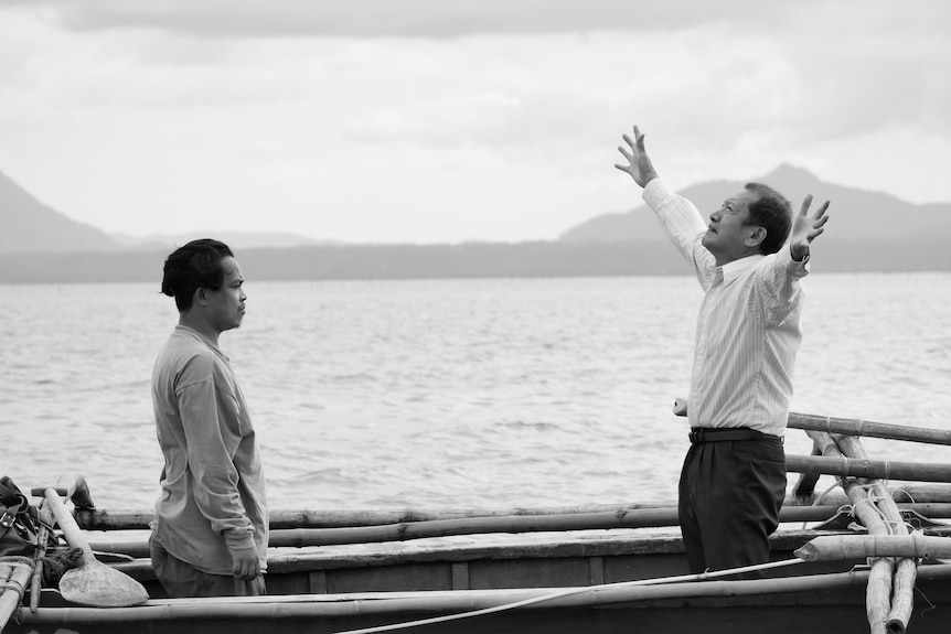 Black and white photo of two Filipino men standing facing each other on a boat, one with arms up to the sky, mountains behind