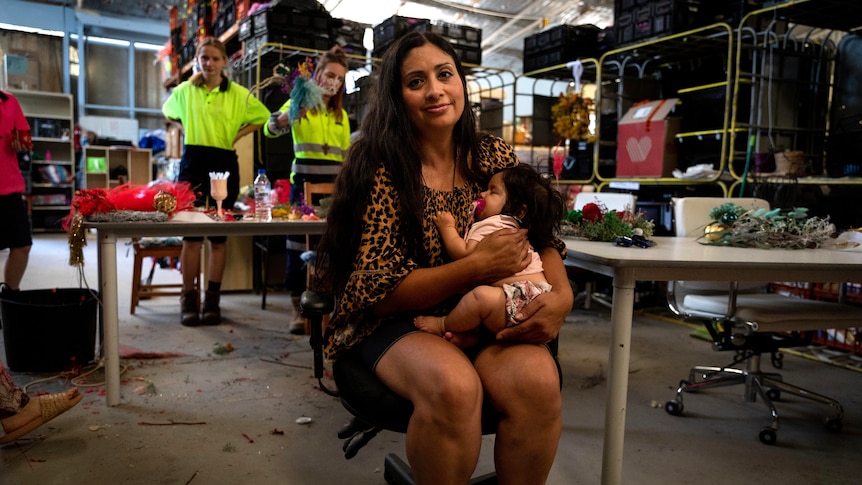 A woman holds a sleeping baby while sitting in a chair in a warehouse, people making Christmas decorations behind her.