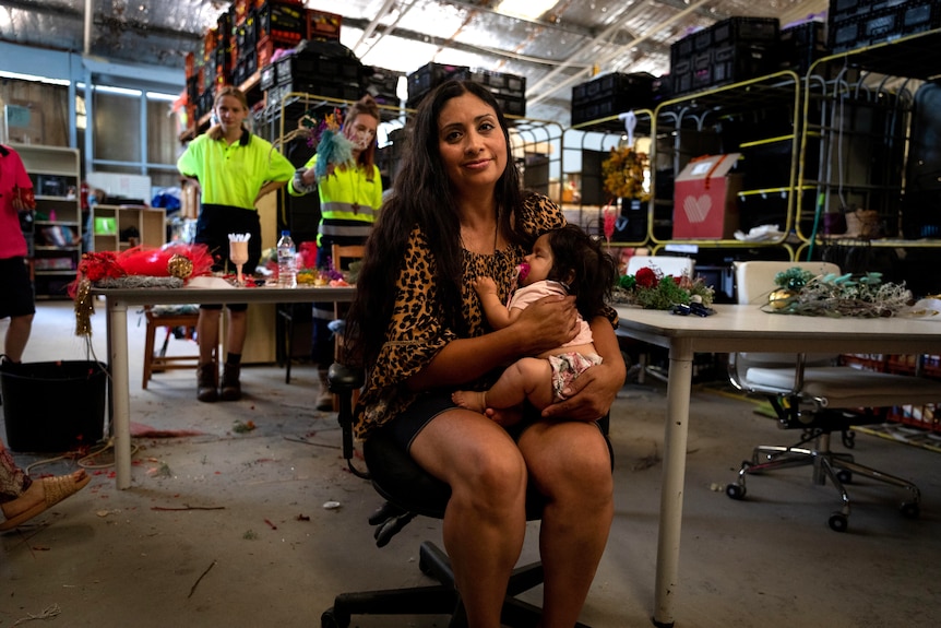 A woman holds a sleeping baby while sitting in a chair in a warehouse, people making Christmas decorations behind her.