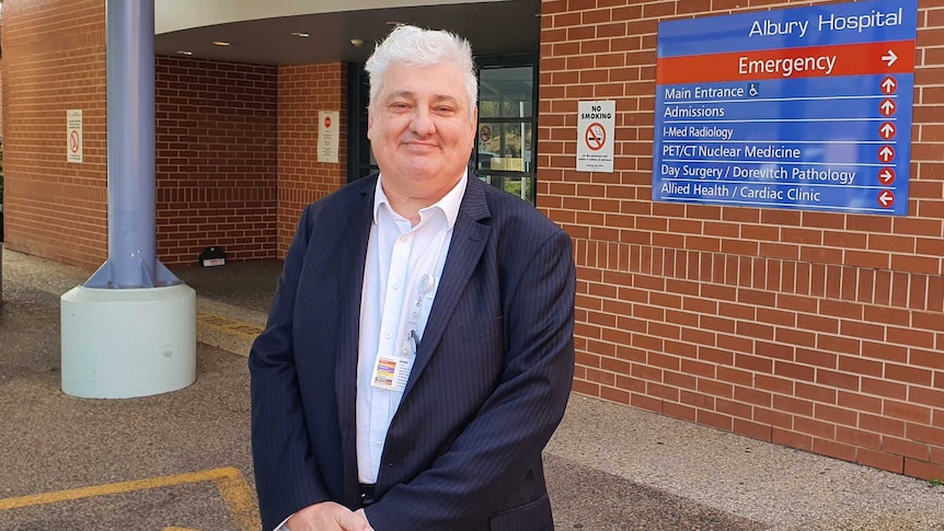 A man with grey hair, dressed in a suit, stands at the entrance to a hospital.