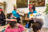 Photo of two women wearing colourful hats and holding books speaking in an animated way with children 
