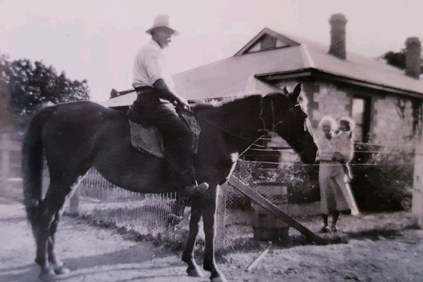 Old black and white photo of a man riding a horse while woman stands nearby with a baby on her hip.