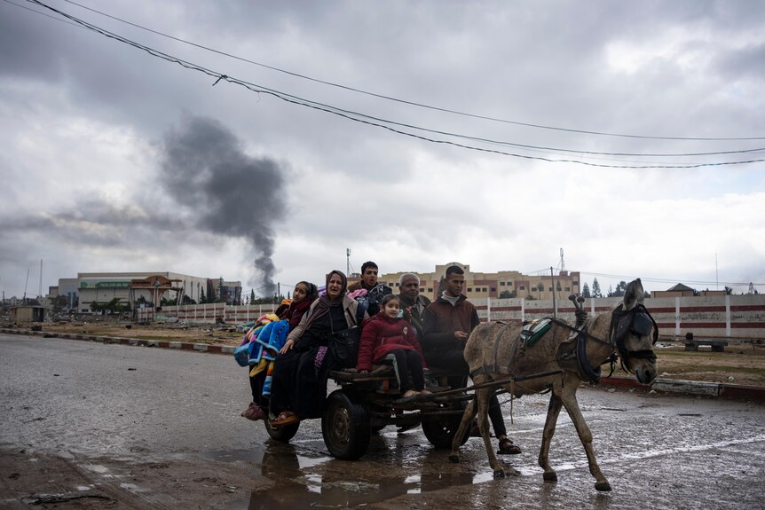 People being carried on a donkey with smoke plumes in the distance 