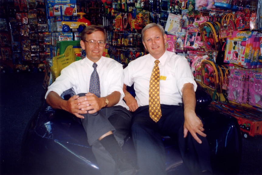 two men are sitting down in a shop with brightly coloured goods behind them, they are wearing a shirt and tie