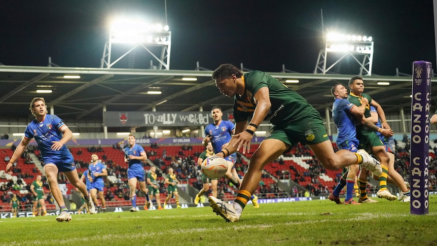 A shot looking across field as an Australian rugby league player runs in to score nearest the camera, and Italian players watch.