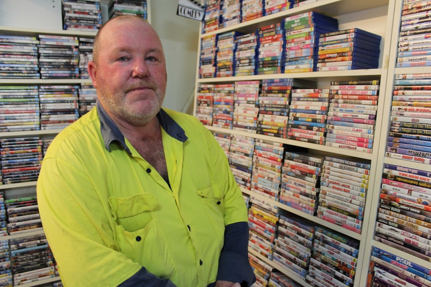 A man in a flouro yellow work shirt standing in front of a wall of DVDs.