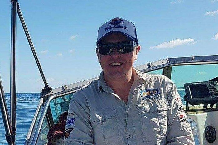 John Craig sits on a boat smiling wearing sunglasses and a cap.