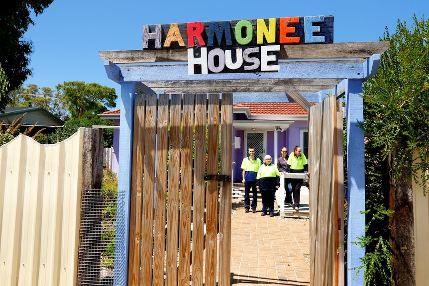 Four people standing inside the gates with a brightly coloured sign which reads 'Harmonee House'.