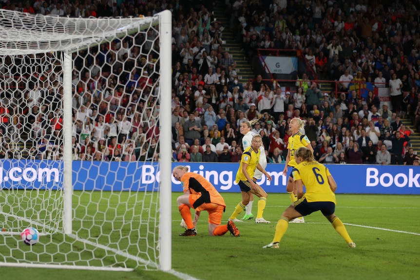 An England women's soccer player sees her backheeled shot go into the net as Swedish keeper and defenders look on.