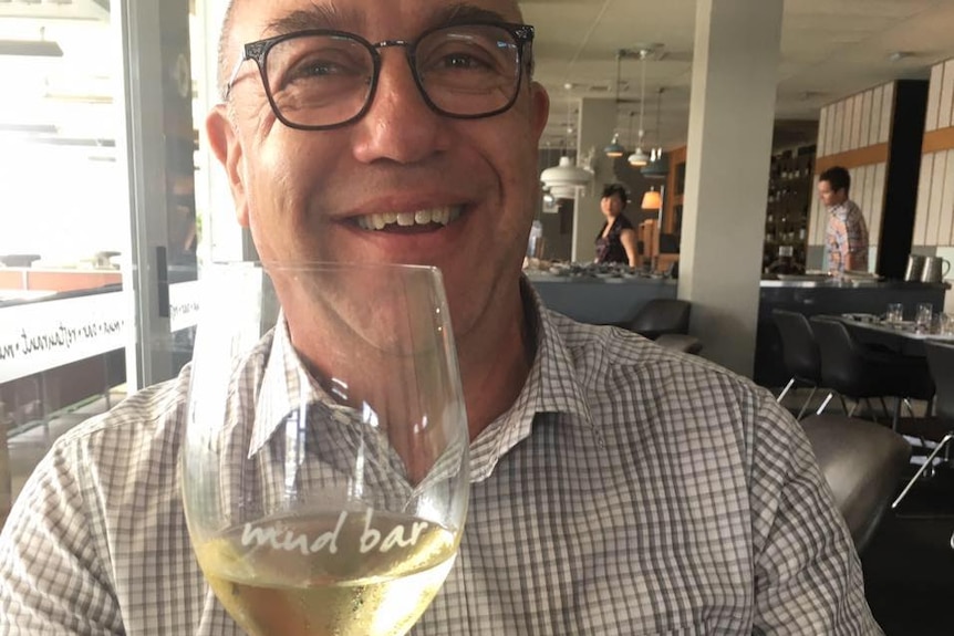 A man wearing glasses smiles at the camera and holds up a glass of wine.