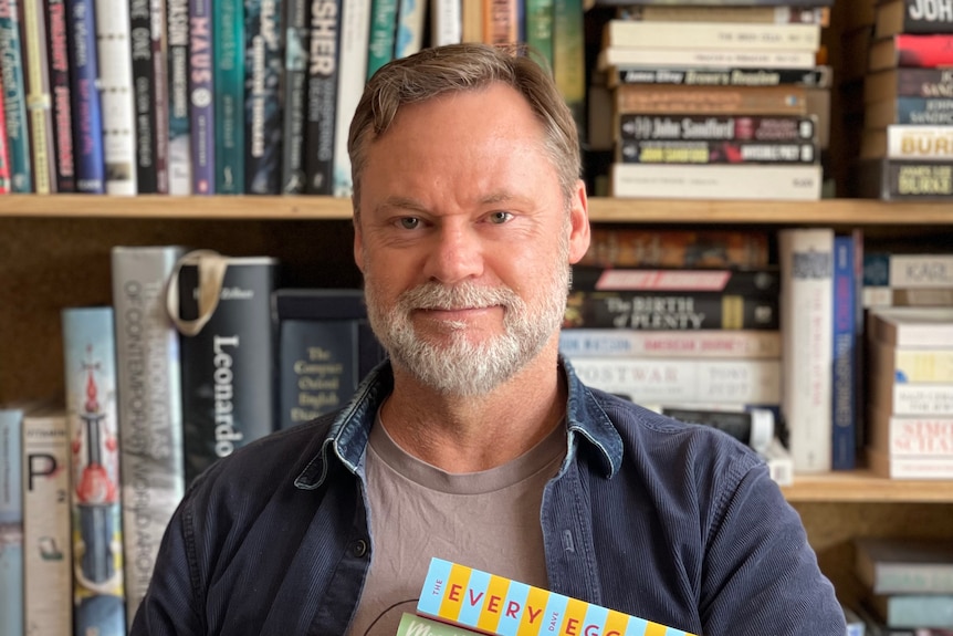 Middle-aged man, grey beard, light brown hair, wears blue jacket, grey tee, smiles, stands in bookshop holds book stack.