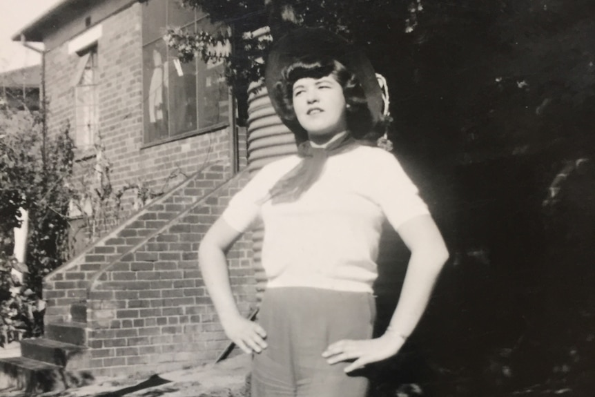 A young woman in the 1940s poses for a photograph in a suburban street in front of a brick house.
