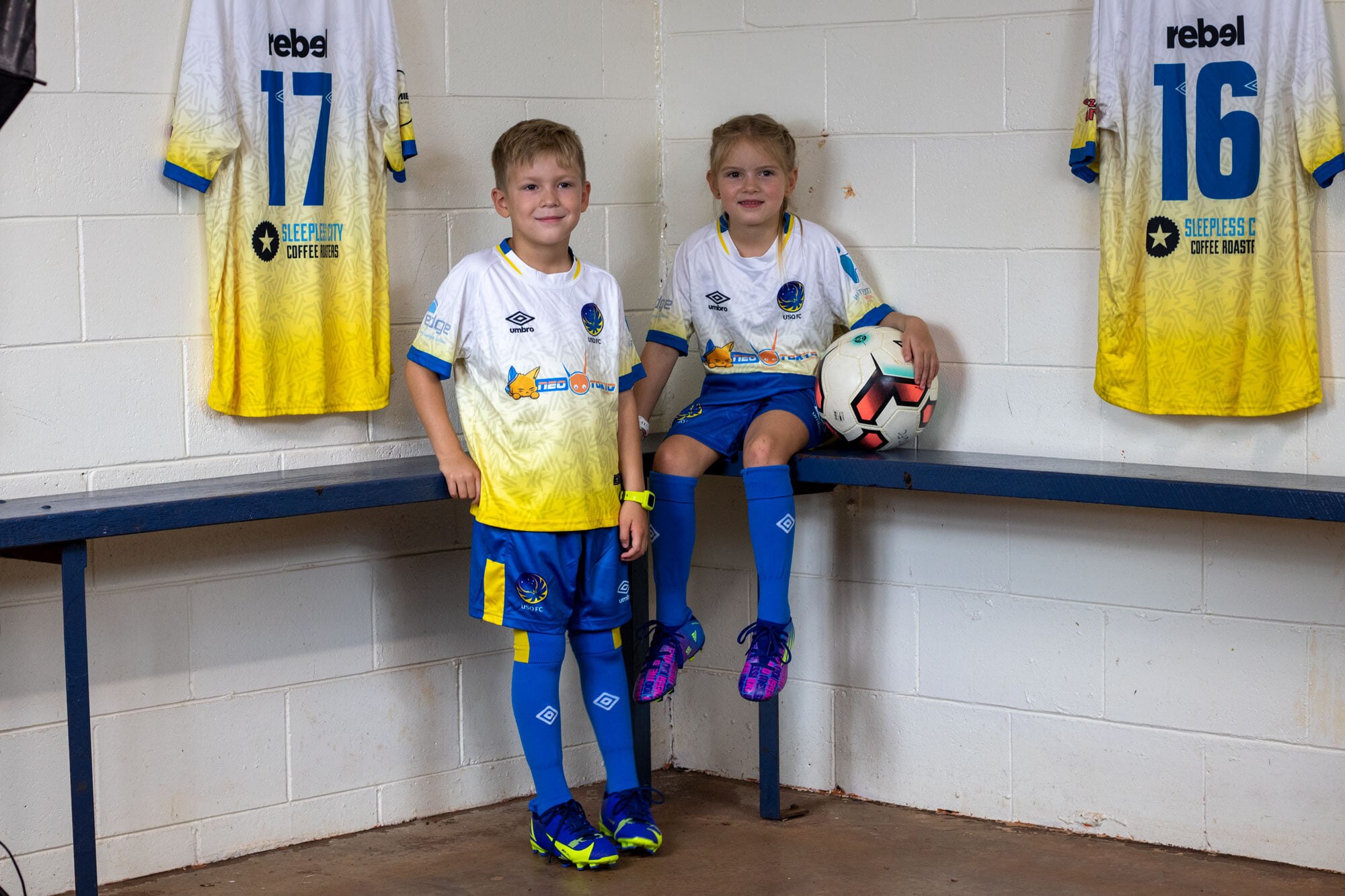 Two kids in soccer uniform one holding a soccer ball smiling at camera. 