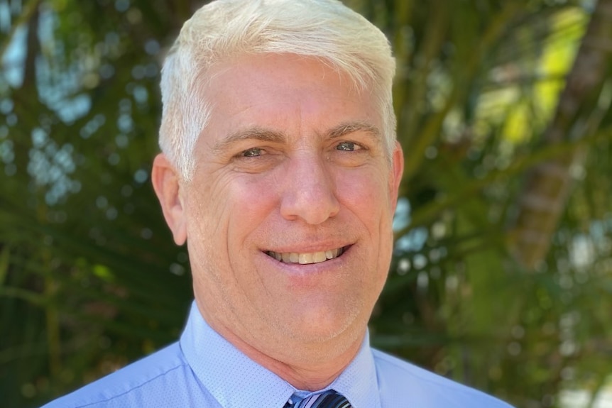 A man with white hair smiles. He is wearing a blue shirt and blue striped tie. There is foliage in the background.