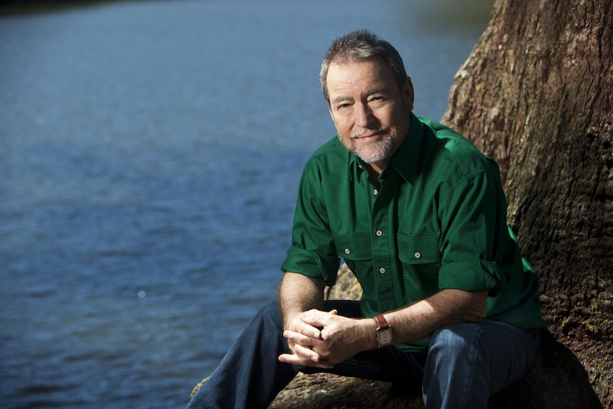 Man in green shirt and blue jeans sits on tree stump next to river