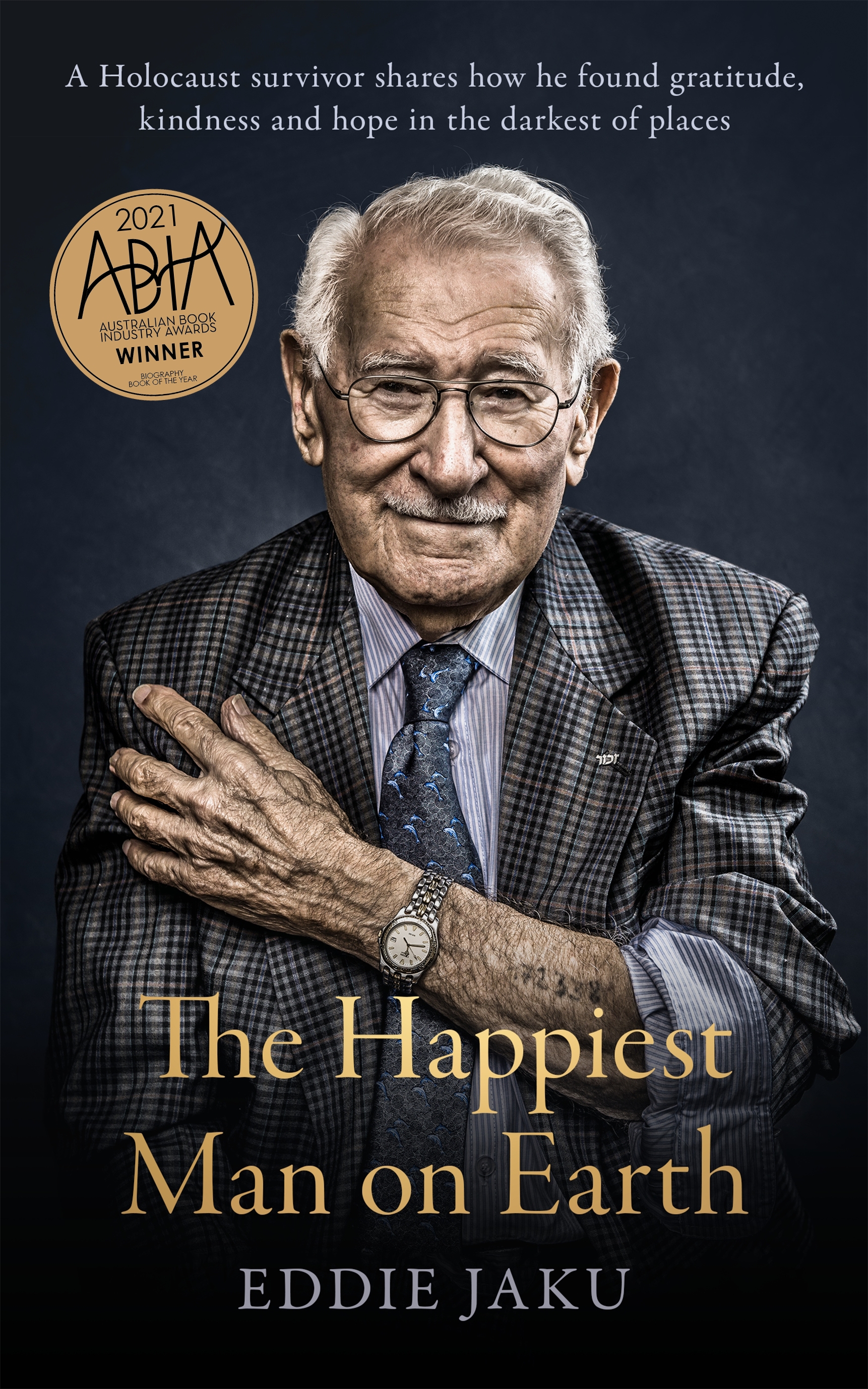 A book cover: an old man with silver hair, glasses and a wry smile wears a suit, one sleeve pushed up showing a tattooed number