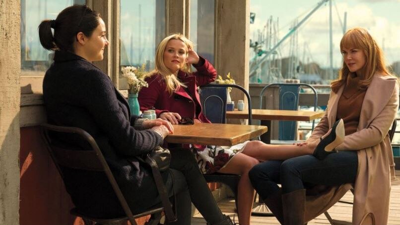 Three women sit around an outside cafe table, one rests her leg on the thigh of another. Ship masts are in the background.