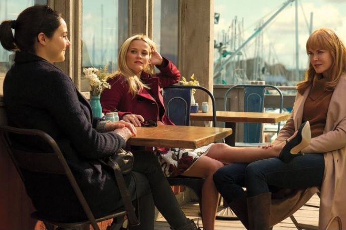 Three women sit around an outside cafe table, one rests her leg on the thigh of another. Ship masts are in the background.
