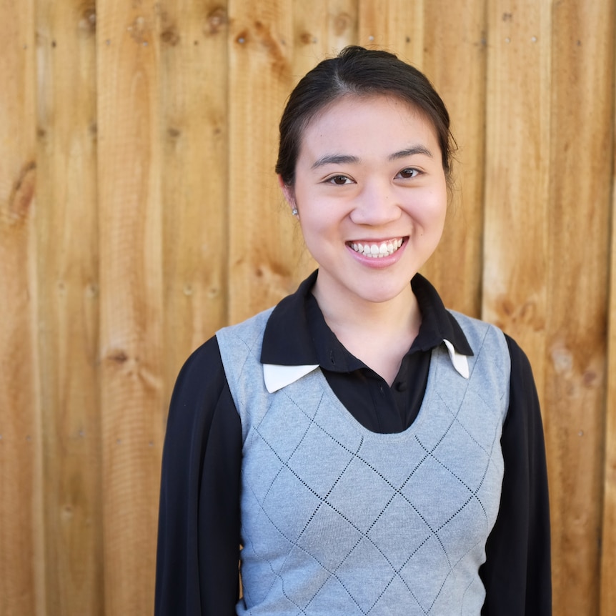 A young smiling woman stands near a panneled timber wall.