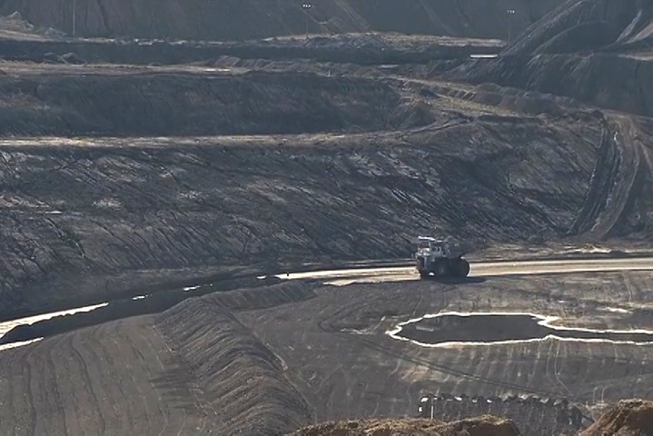 A photo taken from a hill of a large truck moving through a grey mine with puddles lit up by the morning sun.