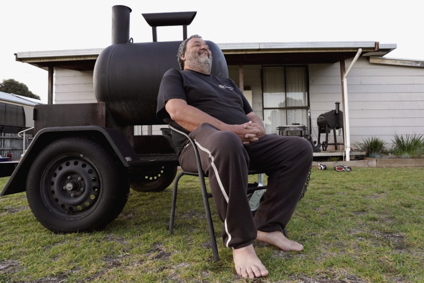 Stuart Thomson sits back in his chair laughing on the front lawn next to his large smoker.