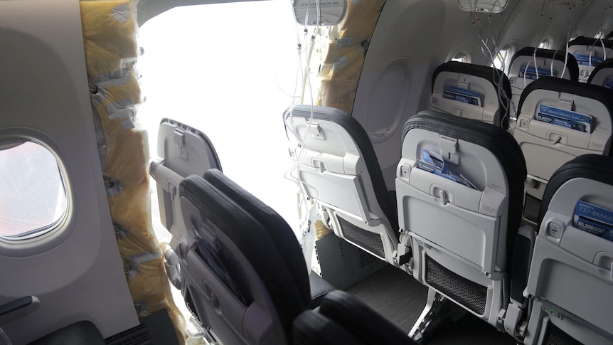 A row of airplane seats sits next to a large hole in the inner wall of a plane.