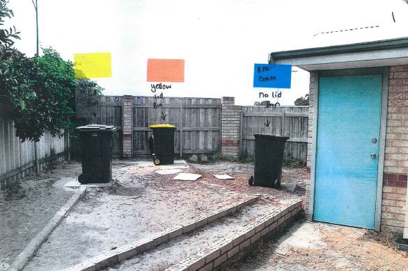 A bare sand backyard with three wheelie bins and evidence stickers on the photo.