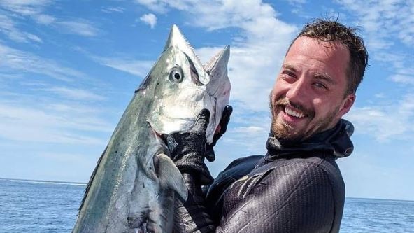 A man in a wet suit holds up a large fish he's caught on a boat in the ocean.