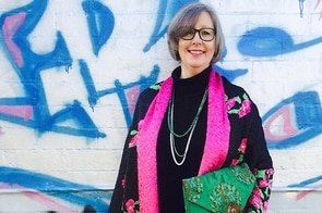 A woman wearing a pink scarf holds a green bag and stands smiling in front of a wall covered in grafiti