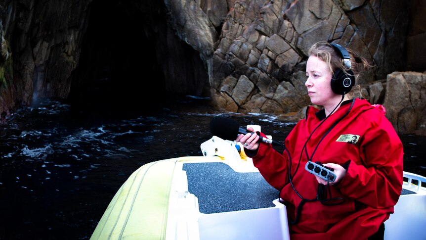 A woman in a red cold-weather jacket captures audio of the ocean lapping against a boat.