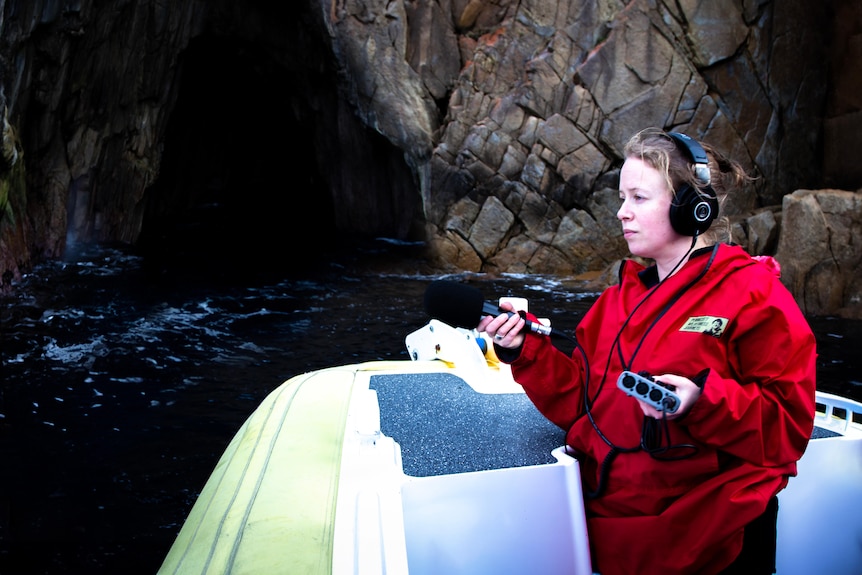 A woman in a red cold-weather jacket captures audio of the ocean lapping against a boat.